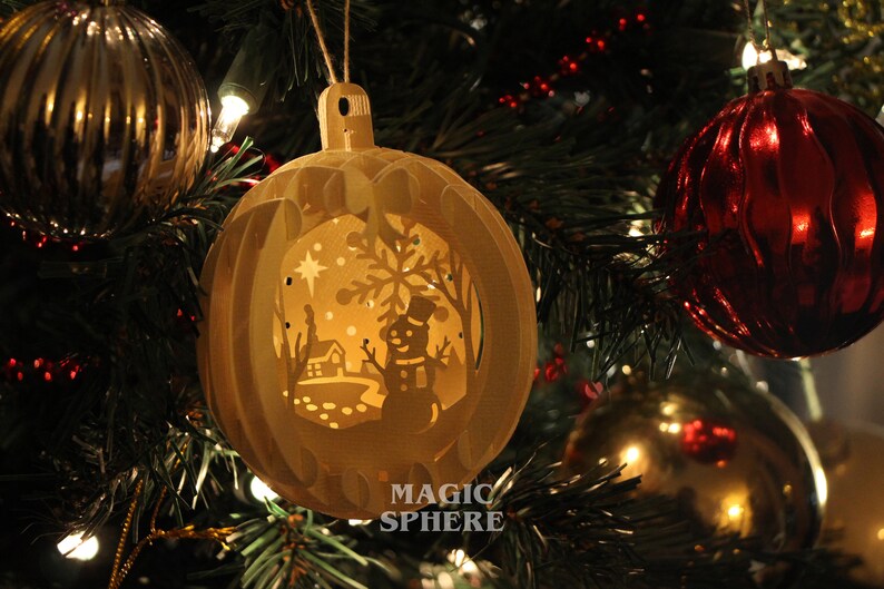 May include: A white, paper Christmas ornament with a cut-out design of a snowman, a house, and a star. The ornament is lit from inside, creating a warm glow. The text "MAGIC SPHERE" is printed on the bottom of the ornament.
