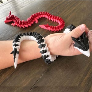 May include: A red and a black and white articulated dragon toy. The black and white dragon is wrapped around a wrist. The red dragon is on a wooden surface. The dragons have a detailed, segmented design.