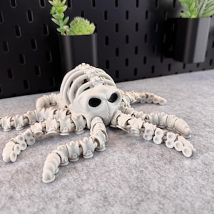 May include: A white, articulated octopus skeleton figurine with a detailed skull and segmented tentacles. The figurine is lying on a gray surface, showcasing its skeletal structure. The octopus is a unique decorative item.