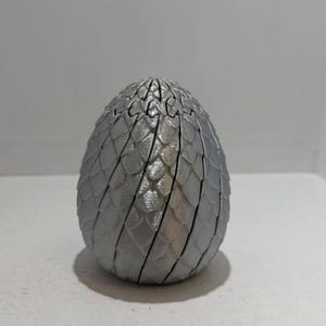 May include: A silver egg-shaped sculpture with a textured, scale-like pattern. The egg stands on a white surface. The sculpture has a segmented design, with lines separating the individual scales. The egg is approximately 15 cm tall.