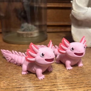 Op de afbeelding: Twee roze axolotl-beeldjes met rode kieuwen en lachende gezichten op een houten oppervlak. De beeldjes zijn gedetailleerd en lijken handgemaakt. De axolotls zijn lichtroze.