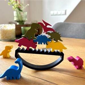 Op de afbeelding: Een kleurrijk houten dinosaurussen balanceerspeelgoed. De set bevat een zwarte wiebelbasis en verschillende felgekleurde dinosaurusfiguren in geel, blauw, roze, groen en rood. Het speelgoed is ontworpen om evenwicht en coördinatie te bevorderen.