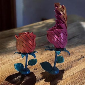 Puede incluir: Dos esculturas decorativas de rosas de metal. Una es de color rojo oscuro, la otra magenta vibrante. Ambas tienen tallos y hojas de color azul verdoso. Los pétalos de rosa tienen una textura acanalada. Se exhiben sobre una superficie de madera.