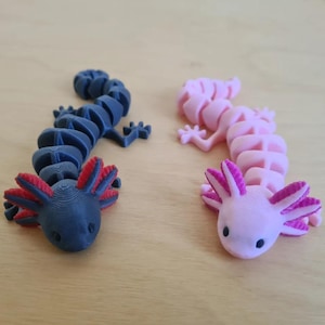 May include: Two articulated axolotl figurines. One is dark blue with red and black gills, the other is pink with purple gills. The axolotls are on a light brown surface. These 3D-printed toys are a unique gift idea.