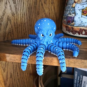 Puede incluir: Una figura de pulpo azul con lunares blancos, colocada sobre una estantería de madera. El pulpo tiene tentáculos articulados y una cabeza detallada con ojos expresivos. Un artículo decorativo para la decoración del hogar.