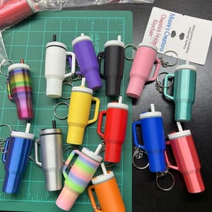 May include: A collection of colorful chapstick holder keychains in various colors, including white, purple, black, pink, teal, blue, silver, yellow, red, orange, and rainbow patterns. Each keychain has a handle and a white lid. The text "Moore's Creations Chapstick Holder Keychain" is visible.