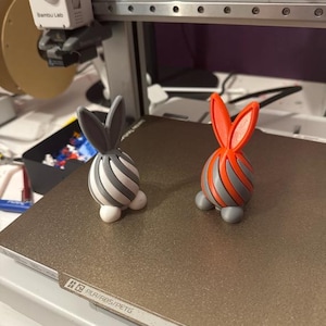 May include: Two 3D-printed bunny figurines. One is gray and white striped with gray ears, the other is orange and gray striped with orange ears. Both have white feet. They are on a brown surface.