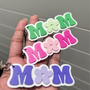 May include: Three keychains spelling "MOM" in green, pink, and purple, each with a flower accent. The letters and flowers are raised, with a white border. Each keychain has a silver clasp.