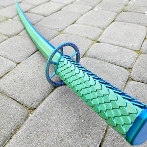 May include: A decorative sword with a curved, green and blue blade. The handle is green with a scale pattern and blue accents. The sword is lying on a gray stone surface.