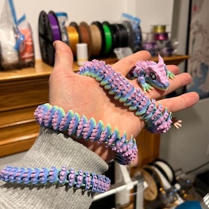 Może przedstawiać: Kolekcja drukowanych w 3D, przegubowych smoków i bransoletek w odcieniach niebieskiego, fioletowego i zielonego. Smok ma szczegółową głowę i ciało, a bransoletki są elastyczne. Przedmioty są trzymane w dłoni, prezentując design i elastyczność.