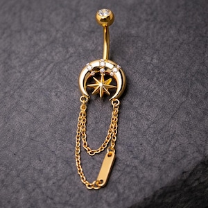 Könnte beinhalten: Goldfarbener Bauchnabelring mit Stern- und Halbmond-Design. Der Ring hat eine herabhängende Kette und einen kleinen, rechteckigen Anhänger. Die Oberseite des Rings hat einen kleinen, runden Edelstein.