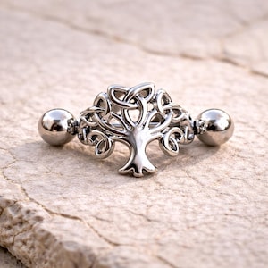 Op de afbeelding: Een zilverkleurige barbell-stijl body piercing sieraden met een Keltisch levensboomontwerp. De sieraden hebben een centraal boommotief met ingewikkeld knoopwerk en twee afgeronde eindballen. Het stuk wordt getoond op een getextureerd, lichtgekleurd oppervlak.