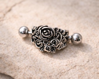 May include: Silver industrial barbell earring with a floral design. The center piece features a cluster of roses and intricate detailing. The barbell has a ball on each end. The jewelry is displayed on a light-colored, textured surface.