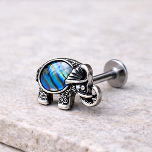 May include: A silver-toned lip piercing featuring an elephant design. The elephant has a blue and green iridescent abalone shell inlay. The piercing has a flat back and a straight post for insertion. The elephant has detailed scrollwork.