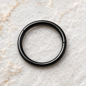 Puede incluir: Un aro circular con un acabado negro liso y pulido. La pieza de joyería presenta un diseño sin costuras con un único punto de bisagra visible, creando una forma circular continua y sencilla adecuada para diversos piercings corporales.