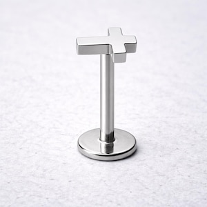 May include: A silver-colored, cross-shaped lip piercing stud. The cross is at the top of a straight post, which is attached to a round base. The jewelry is made of a shiny, metallic material and is set against a light background.