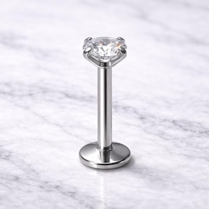 May include: A silver-colored labret stud with a clear, round gemstone. The stud has a flat base and a long, slender post that leads to the gemstone setting. The gemstone is held in place by four prongs.