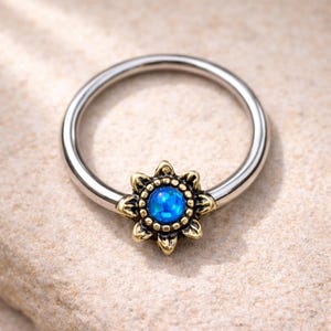 Può includere: Un anello per il naso color argento con un design a fiore color oro e una pietra centrale blu. Il fiore ha un design dettagliato dei petali. L'anello è posto su uno sfondo neutro.