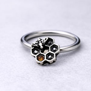 Pode incluir: Anel de prata com design de favo de mel, uma pequena abelha e uma pedra âmbar. O anel tem uma banda polida e um padrão de favo de mel texturizado e detalhado, para os amantes da natureza.