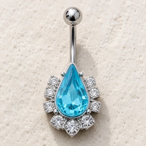 May include: A silver-toned belly button ring featuring a central pear-shaped light blue gemstone. The teardrop-shaped stone is surrounded by a halo of small, clear, round-cut white crystals set in a metallic frame with a smooth top ball.