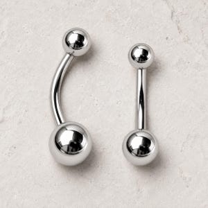 May include: Two silver-toned body jewelry pieces. One is a curved barbell with a ball at each end, and the other is a straight barbell with a ball at each end. Both pieces feature a polished, reflective metallic finish with smooth, rounded spheres.