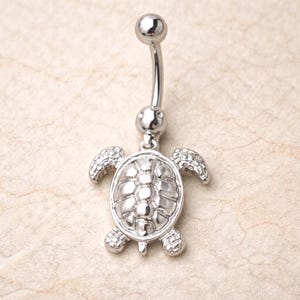 May include: A silver-colored belly button ring featuring a turtle design. The ring has a curved barbell with a small ball at the top and a turtle pendant at the bottom. The turtle has a detailed shell and flippers.