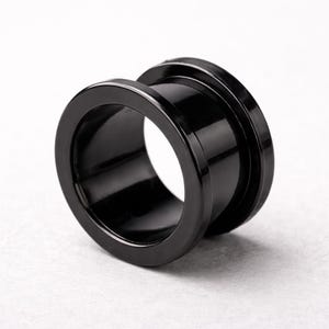 Könnte beinhalten: Ein schwarzer Ohr-Tunnel, ein Schmuckstück zur Körpermodifikation. Der Tunnel besteht aus glänzendem Material und hat eine zylindrische Form mit einem großen, offenen Zentrum. Er dient zur Dehnung des Ohrläppchens.