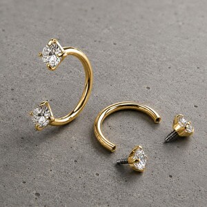 May include: Two gold circular barbell body jewelry pieces featuring clear round-cut gemstones at each end. One piece is fully assembled, while the other is shown disassembled with two separate gemstone attachments with threaded metal posts.