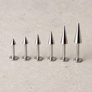 Op de afbeelding: Een set van zes zilverkleurige metalen labret-piercings, gerangschikt op hoogte. Elk sieraad heeft een platte ronde basis, een gladde cilindrische staaf en een puntige kegelvormige top, afgewerkt met een glanzende, metallic uitstraling.