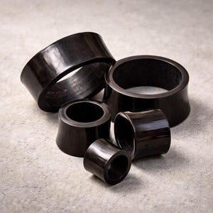 Könnte beinhalten: Fünf schwarze Ohr-Dehnstäbe in verschiedenen Größen, hergestellt aus einem dunklen, glänzenden Material. Die ausgestellten Dehnstäbe werden auf einer hellgrauen, strukturierten Oberfläche präsentiert. Die Ohr-Dehnstäbe haben eine glatte, polierte Oberfläche. Der größte hat einen Durchmesser von ca. 2,5 cm.