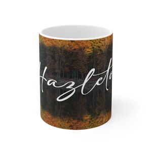 Puede incluir: Taza de cerámica blanca con una foto de un bosque reflejándose en un lago. El texto "Hazleton" está escrito en cursiva blanca en la taza.