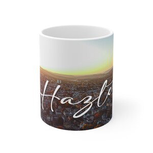 Puede incluir: Taza de cerámica blanca con una fotografía de una ciudad al atardecer. El texto "Hazleton" está escrito en cursiva blanca en la taza.