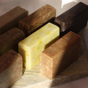 May include: Assortment of six handmade soap bars in various shades of brown and yellow. The bars are arranged on a light brown stone surface.