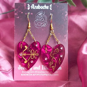 May include: A pair of heart-shaped earrings with gold-colored chains and hooks. The earrings are a deep pink color with gold-colored designs and clear gems. The earrings are displayed on a pink satin background.