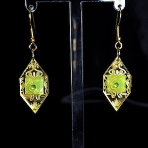 May include: Gold-coloured earrings with a geometric design. Each earring has a central square of light green with a swirling pattern, framed by a gold filigree border. The earrings hang from gold-coloured hooks.