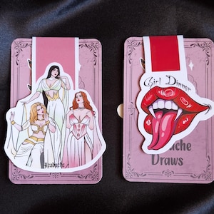 May include: Two pink bookmarks. One shows three women in white and gold dresses, the other has a red mouth with fangs and the text "Girl Dinner". Both have a decorative pink border and a red tab.