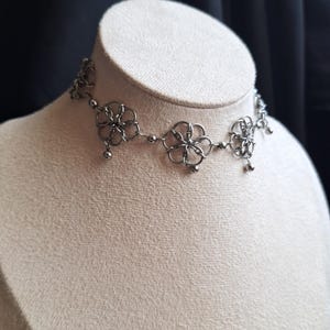 Peut inclure: Un collier ras du cou argenté avec un motif floral. Le collier présente des formes de fleurs ajourées reliées par de petites perles et des breloques sphériques pendantes. Le bijou est exposé sur un mannequin.