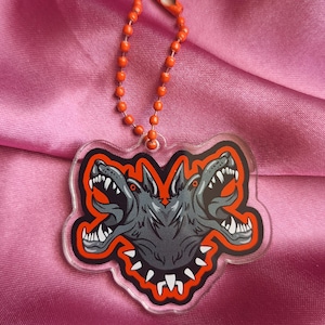 May include: A clear acrylic keychain featuring a two-headed dog design with sharp teeth and an open mouth. The dog is gray with an orange outline and a matching orange ball chain. The keychain is on a pink satin background.