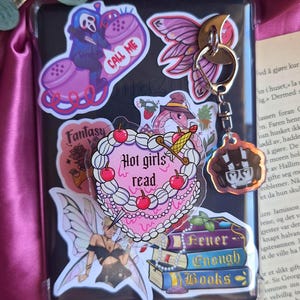 Könnte beinhalten: Eine schwarze E-Reader-Hülle mit Aufklebern zum Thema Fantasy, darunter eine Fee, ein herzförmiger Kuchen mit dem Text "Hot girls read" und ein Schlüsselanhänger mit Totenkopf-Design. Der Hintergrund ist ein Satin-Stoff.