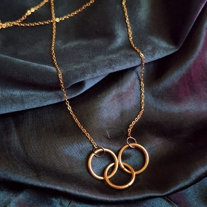 May include: A gold chain necklace with three interlocking rings as a pendant. The necklace is on a dark blue fabric background.