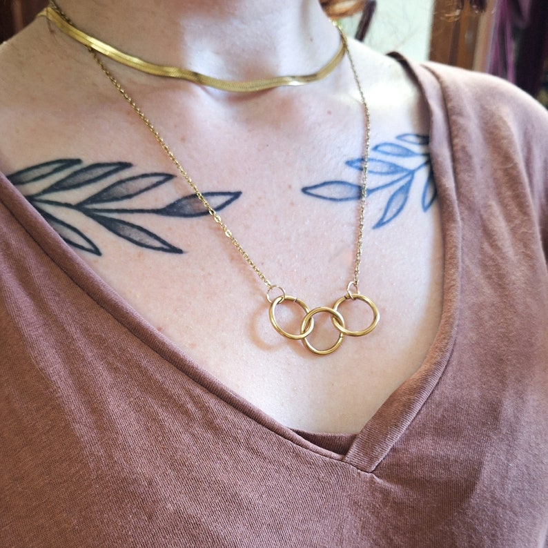 May include: A gold chain necklace with three interlocking circles pendant. The necklace is layered over a thin gold chain.