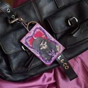 Puede incluir: Un bolso de cuero negro con una etiqueta decorativa que presenta una calavera con capucha, mariposas y el texto "Hey!". La etiqueta está sujeta a la bolsa con una cadena dorada y una etiqueta de cuero negro que dice "L & CO".