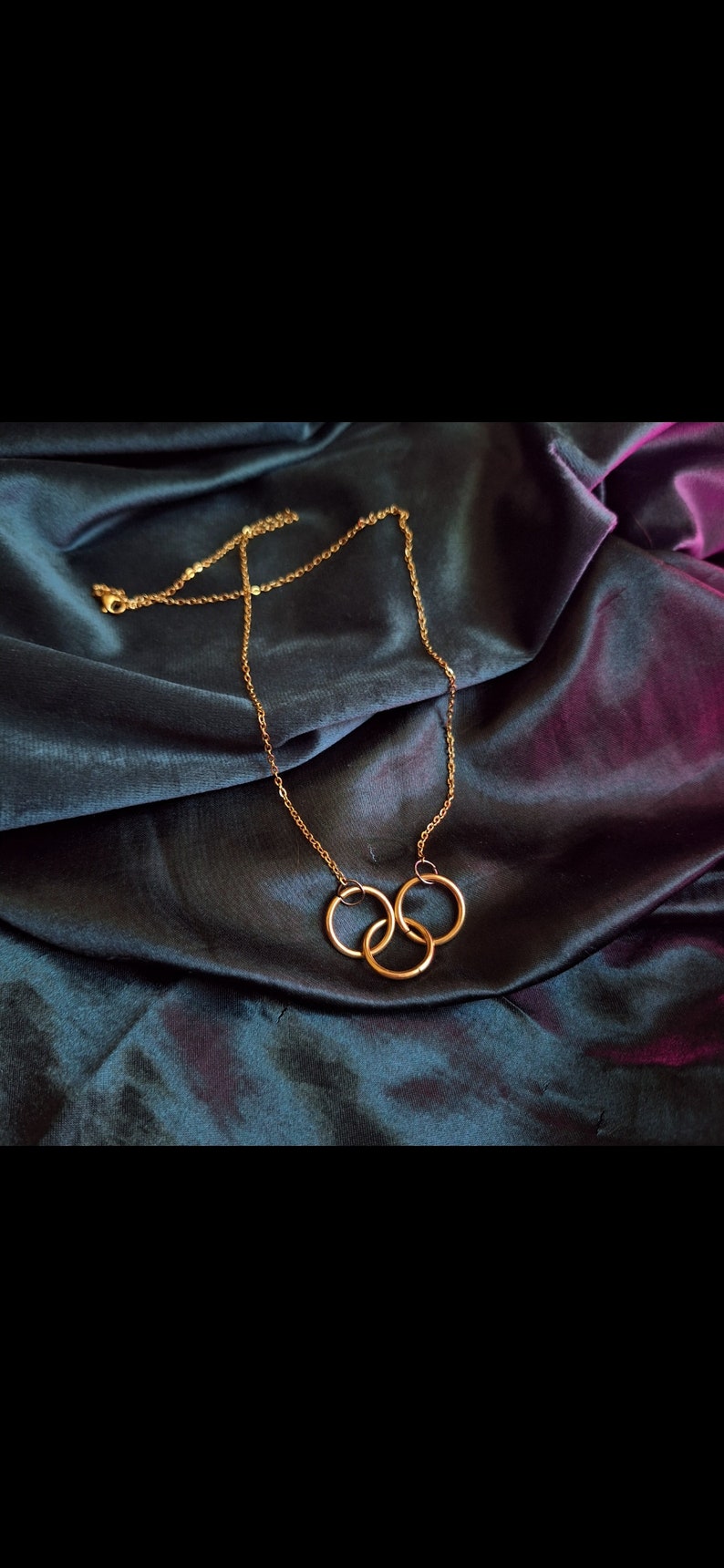 May include: A gold chain necklace with three interlocking rings as a pendant. The necklace is on a dark blue fabric background.