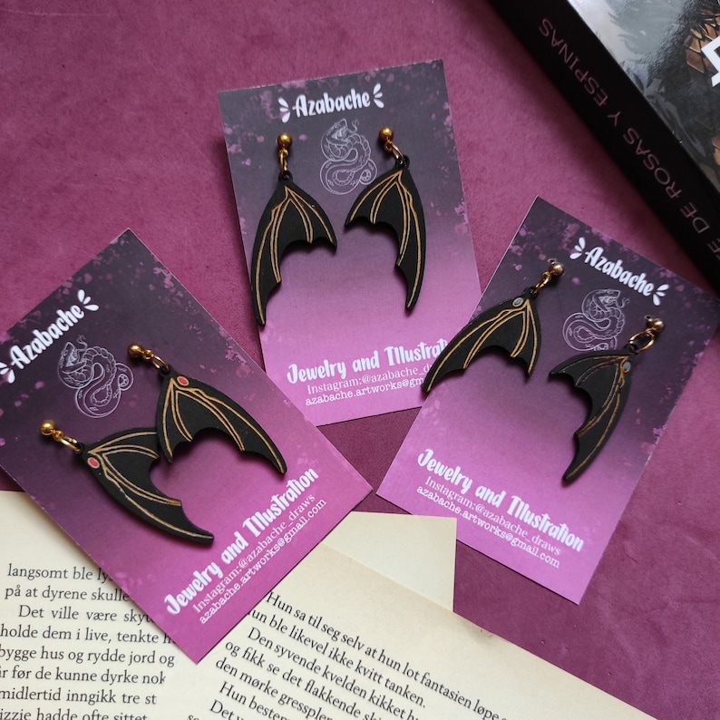 May include: A pair of black bat wing earrings with gold accents. The earrings are displayed on a white card with the text "Azabache Jewelry and Illustration" and social media information.