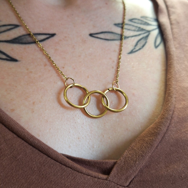 May include: A gold necklace with three interlocking rings. The necklace is worn by a person with a tattoo on their chest.