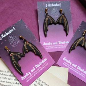 May include: A pair of black bat wing earrings with gold accents and red accents on the wings. The earrings are displayed on a card with the text "Azabache Jewelry and Illustration" and the Instagram handle "@azabache_draws".