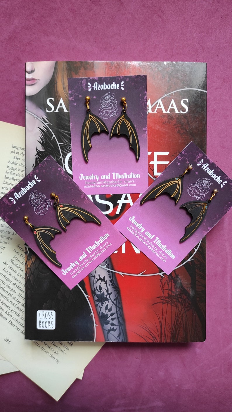 May include: A pair of black and gold bat wing earrings on a pink and purple background. The earrings are on a card with the text "Azabache" and "Jewelry and Illustration" with contact information.