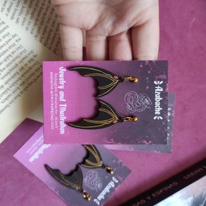 May include: Black and gold bat wing earrings with red accents. The earrings are on a card with the text "Azabache" and "Jewelry and Illustration".