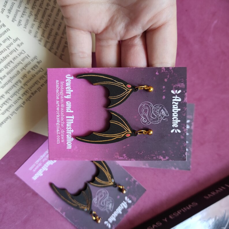 May include: Black and gold bat wing earrings with red accents. The earrings are on a card with the text "Azabache" and "Jewelry and Illustration".