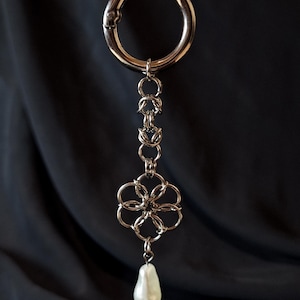 Könnte beinhalten: Ein silberfarbener Schlüsselanhänger mit einem großen kreisförmigen Verschluss, verbunden mit einer Kette mit floralem Design und einer tropfenförmigen Perle. Der Hintergrund ist ein dunkler, drapierten Stoff.